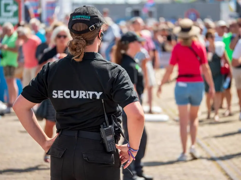 A security guard monitoring a crowded outdoor event, wearing a black uniform and radio.