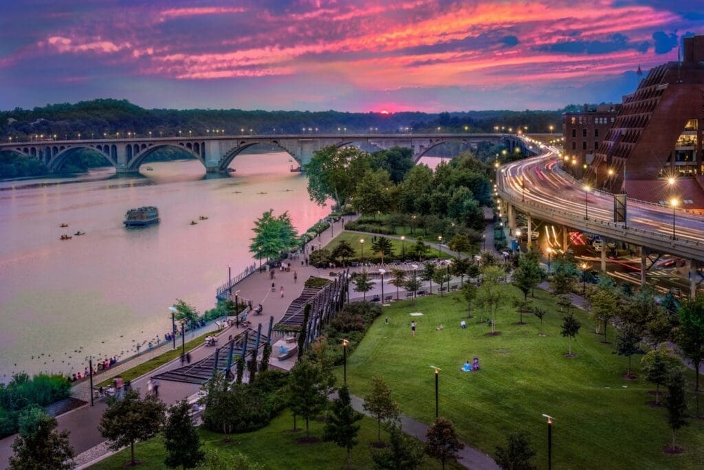 Sunset view of the Georgetown Waterfront Park along the Potomac River, with the Key Bridge, city lights, green parkland, and people relaxing by the river in Washington, DC.