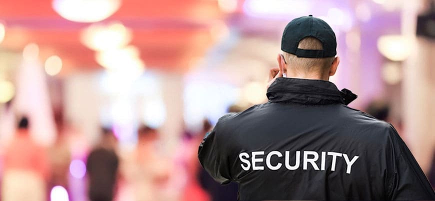 Security guard wearing a jacket labeled “SECURITY” monitoring a crowd at an indoor event venue.
